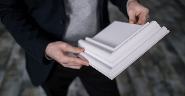 Person holding a white decorative cornice or molding sample, wearing a dark blazer and gray sweater.