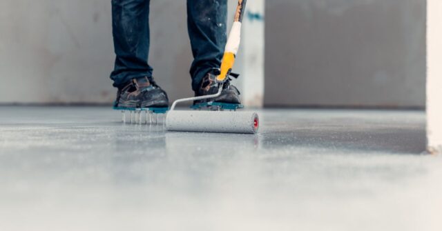 Close-up of a worker wearing spiked shoes using a paint roller to apply a coating to a smooth concrete floor during renovation