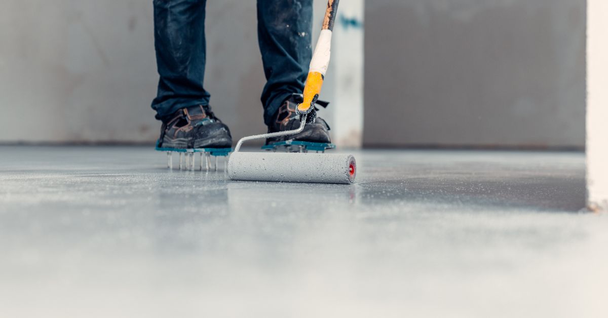 Close-up of a worker wearing spiked shoes using a paint roller to apply a coating to a smooth concrete floor during renovation