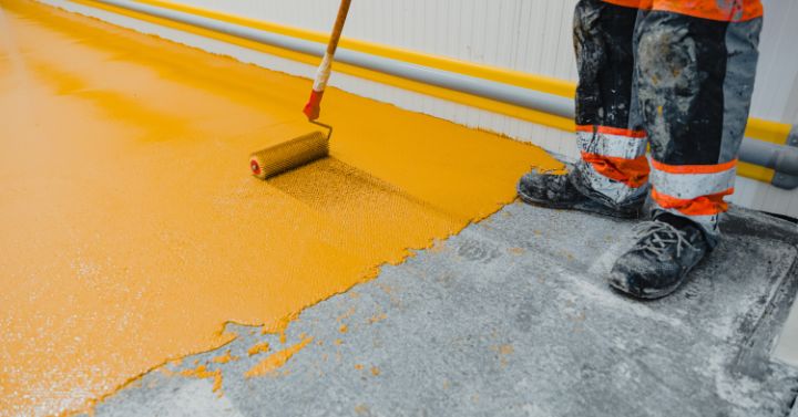 Worker wearing protective boots and clothing using a paint roller to apply bright yellow floor coating along an industrial wall.