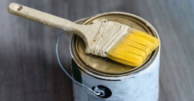 A close-up view of a yellow-bristled paintbrush resting on top of a sealed paint can, placed on a dark wooden floor.