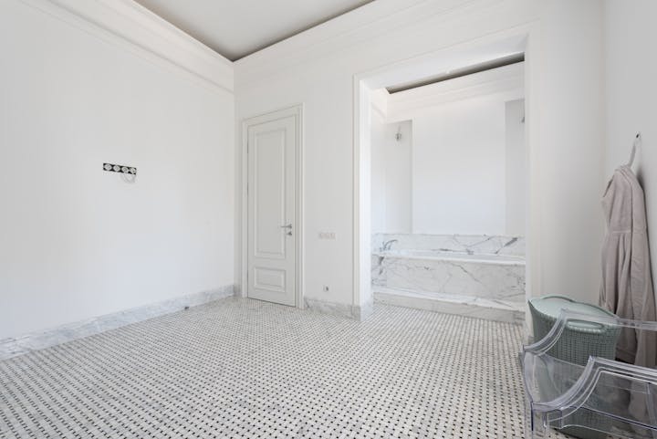 Bright white bathroom interior with marble flooring and baseboards, a door, and elegant wall cornices.