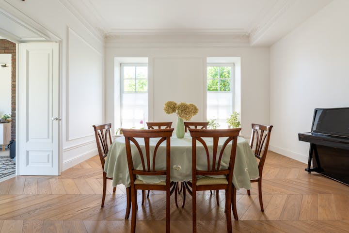 Bright dining room with a round table covered with a light green tablecloth, six wooden chairs, a vase with flowers, and elegant wall cornices.