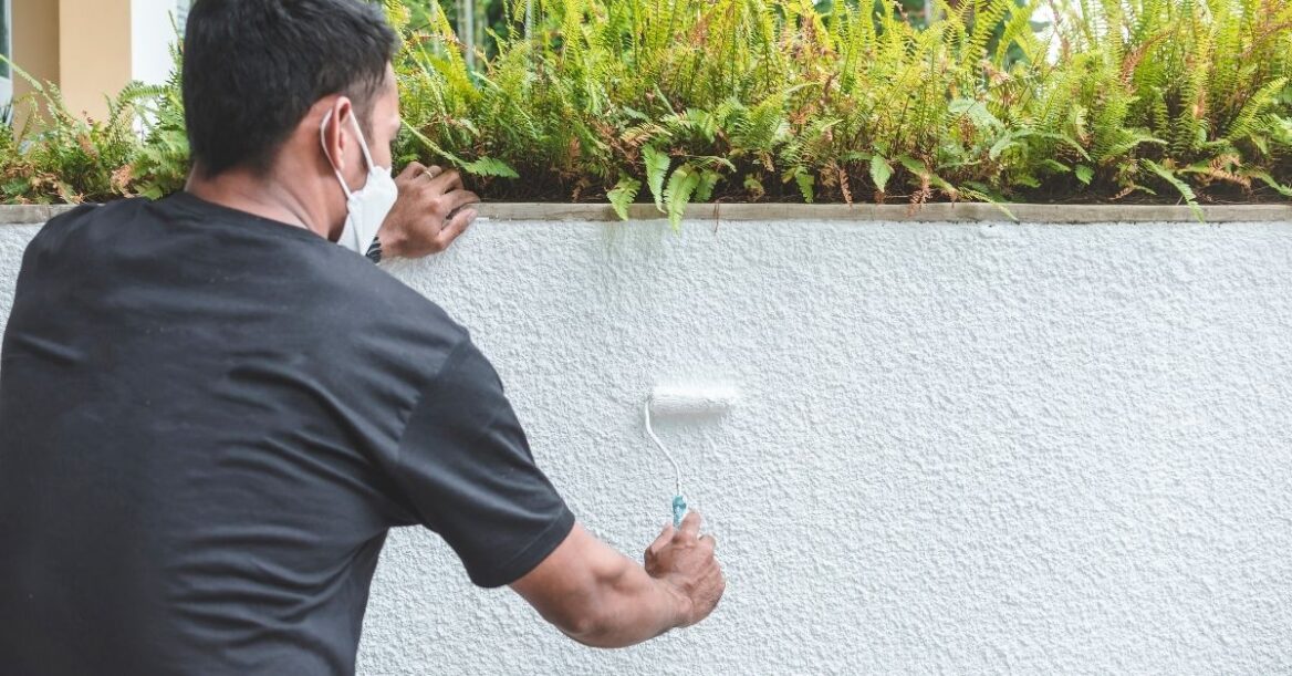 A handyman using a small paint roller to apply white colored epoxy-based paint on the rough textured wall of a ledge near the garage.