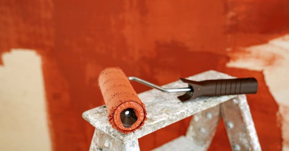 A paint roller coated in reddish-orange paint rests on a stepladder in front of a partially painted wall, suggesting an ongoing home repainting project.