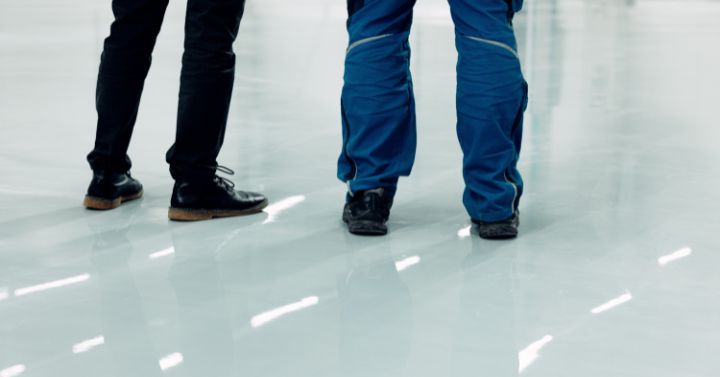 Two people standing on a glossy finished floor, one wearing workwear and the other in dress shoes, suggesting inspection or approval of the surface.
