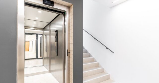 Open stainless steel elevator next to a white staircase in a clean, modern building hallway with bright lighting.