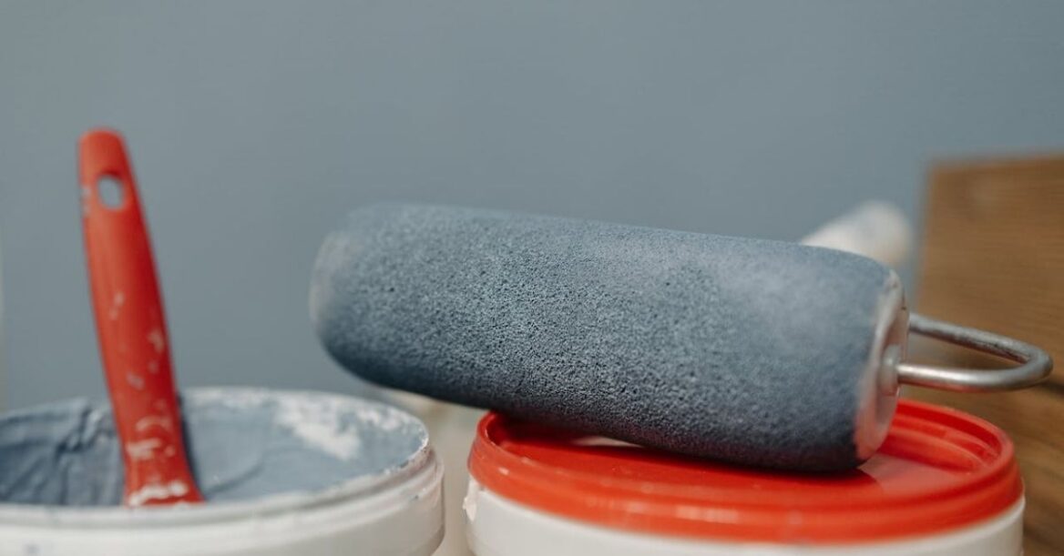 A close-up of a paint roller resting on a red-lidded paint pail, with another open pail of blue-gray paint and a red-handled brush nearby.