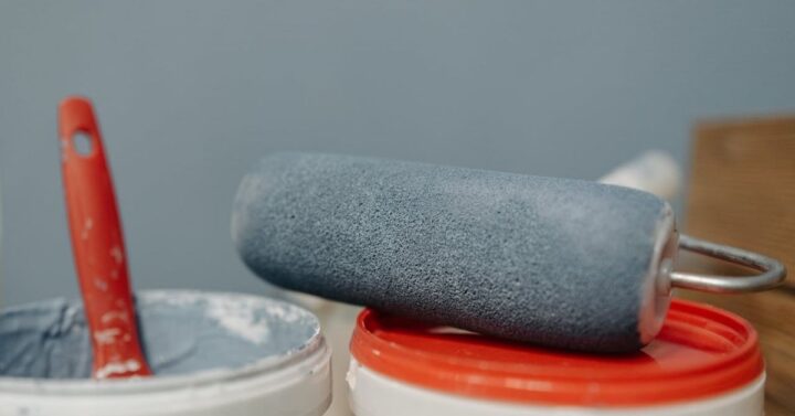 A close-up of a paint roller resting on a red-lidded paint pail, with another open pail of blue-gray paint and a red-handled brush nearby.