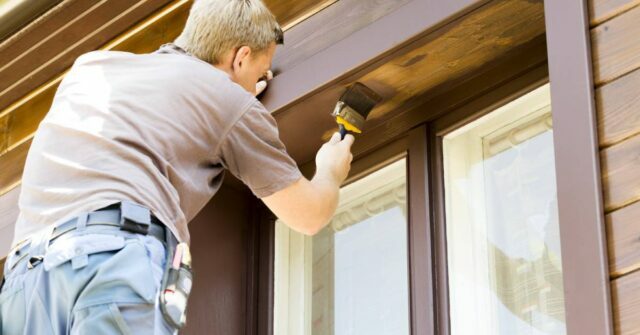 Man with brush painting a wooden house exterior.