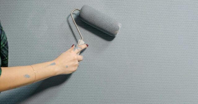 Person painting a textured wall with a gray paint roller, with some paint marks on their hand and arm.