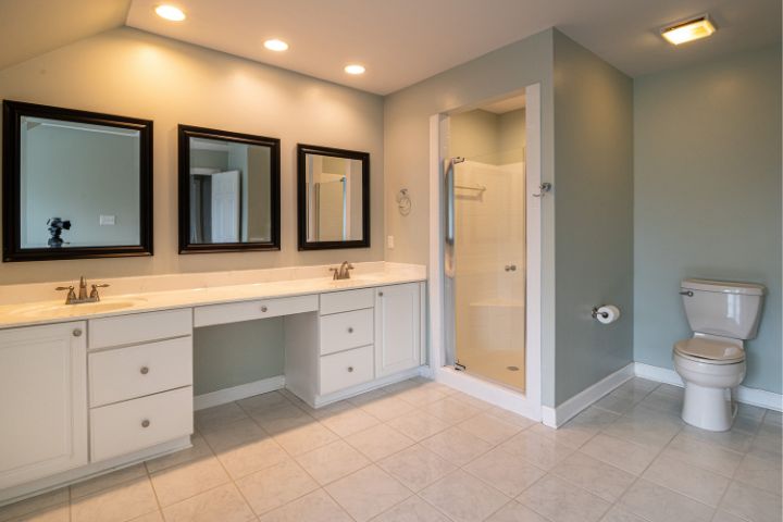 A spacious bathroom featuring a double-sink vanity with three mirrors, a glass-enclosed shower, and a toilet, all set against soft green walls and a tiled floor.