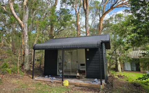 A small black-painted cabin in a forested area with a corrugated metal roof and sliding glass doors after renovation.