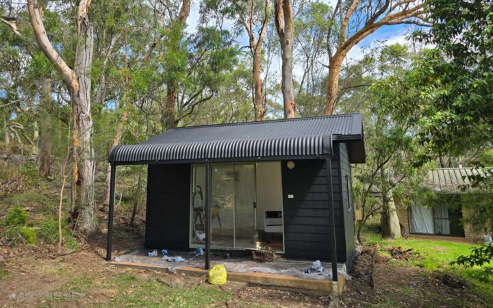 A small black-painted cabin in a forested area with a corrugated metal roof and sliding glass doors after renovation.