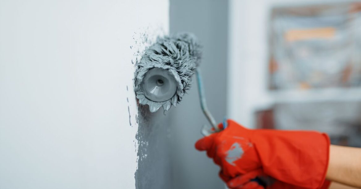 Woman holding paint roller with red gloves, painting a white wall with grey paint