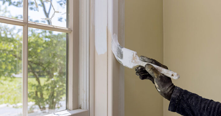 A painter is painting the window trim with a paintbrush using white paint.