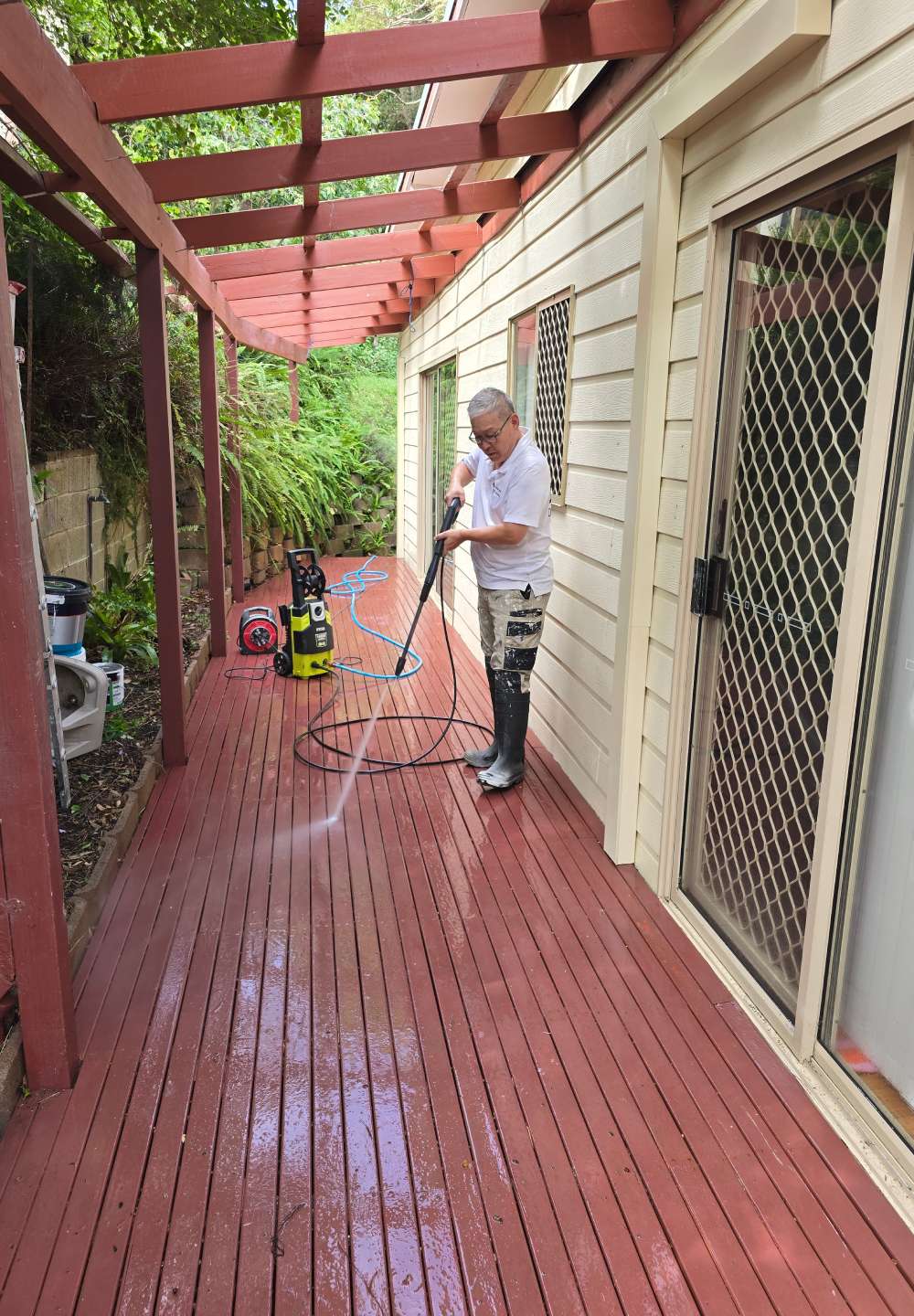 A professional contractor using a pressure washer to clean a red wooden deck alongside a cream-colored house with a pergola overhead.